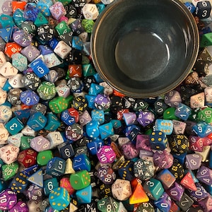 May include: A large bowl filled with a variety of colorful dice, including red, blue, green, yellow, and purple. The dice are scattered randomly in the bowl, creating a visually appealing and chaotic scene.