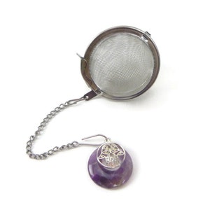 May include: A silver tea infuser with a chain and a purple amethyst charm with a silver Hamsa hand design.