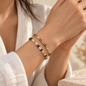 Könnte beinhalten: Zwei Perlenarmbänder am Handgelenk. Ein Armband zeigt ein sich wiederholendes Muster aus quadratischen Perlen in Weiß-, Beige- und Schwarztönen. Das zweite Armband besteht aus quadratischen Perlen in verschiedenen Braun- und Goldtönen.