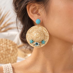 May include: A dangle earring featuring a miniature woven straw hat charm. The hat is decorated with two small turquoise starfish and a central white and blue seashell. A textured turquoise circular stud connects the hat to the earlobe.
