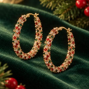 May include: A pair of gold hoop earrings featuring a pavé setting with alternating small round red, green, and clear white stones. The earrings have a hinged post closure mechanism and a circular shape, showcasing a dense arrangement of colorful gems.