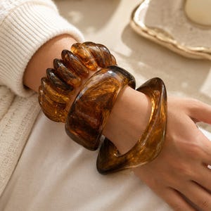 Peut inclure: Un ensemble de trois bracelets en résine brune translucide avec un motif marbré. Un bracelet présente un design côtelé en spirale, tandis que les deux autres sont des joncs larges et lisses aux bords arrondis, portés ensemble au poignet.