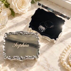 May include: A rectangular silver-toned metal jewellery box with an ornate, scalloped edge and a reflective lid engraved with the name Sophia. The interior is lined with black velvet and contains two silver rings, one featuring a large clear stone.