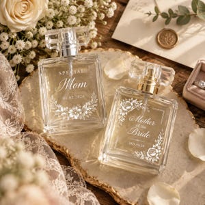 Peut inclure: Deux flacons de parfum en verre transparent avec des vaporisateurs argentés. L'un est gravé avec "SPECIAL Mom 02.02.2026" et une couronne florale. L'autre porte l'inscription "Mother of the Bride 2026.06.28" avec un cadre floral.