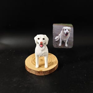 Op de afbeelding: Een wit hondenbeeldje met een roze tong en donkere ogen, staand op een ronde houten schijf als voet. Naast het beeldje staat een kleine rechthoekige foto van een witte hond die op een grindpad zit, passend bij de houding van het beeldje.