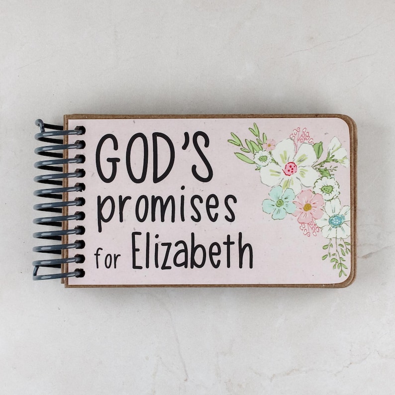 May include: A spiral-bound notebook with the text "GOD'S promises for Elizabeth" in black. The cover is light pink with a floral design in the upper right corner. The notebook has a brown border.