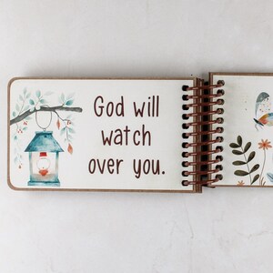 Puede incluir: Un cuaderno de espiral con una p&aacute;gina blanca que presenta el texto marr&oacute;n 'God will watch over you.' A la izquierda hay una ilustraci&oacute;n en acuarela de una linterna azul colgada de una rama con hojas. La siguiente p&aacute;gina muestra una mariposa y flores.