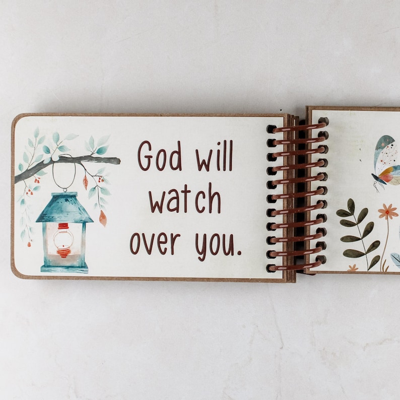 Puede incluir: Un cuaderno de espiral con una p&aacute;gina blanca que presenta el texto marr&oacute;n 'God will watch over you.' A la izquierda hay una ilustraci&oacute;n en acuarela de una linterna azul colgada de una rama con hojas. La siguiente p&aacute;gina muestra una mariposa y flores.