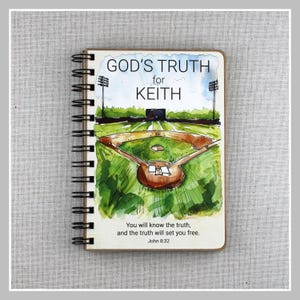 May include: A notebook with a baseball field illustration and the text "GOD'S TRUTH for KEITH". The notebook has a wooden cover and black spiral binding. The bottom of the cover has the text "You will know the truth, and the truth will set you free. John 8:32."