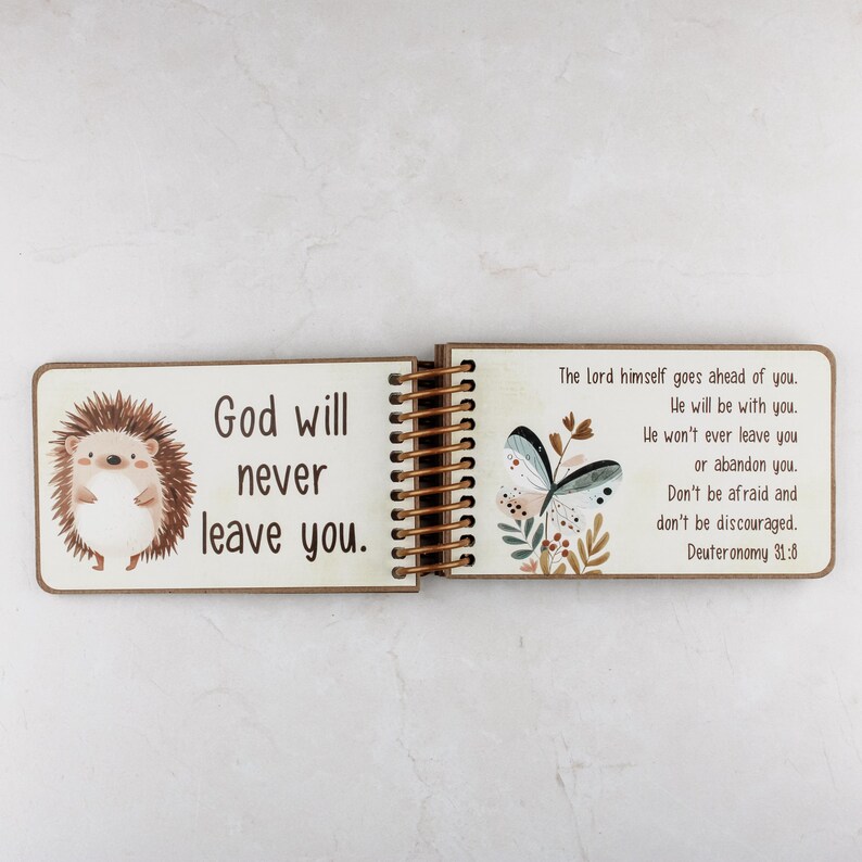 Puede incluir: Un libro de madera con encuadernaci&oacute;n en espiral que presenta un erizo de dibujos animados en la p&aacute;gina izquierda con el texto God will never leave you. La p&aacute;gina derecha muestra una ilustraci&oacute;n de mariposa y el texto The Lord himself.