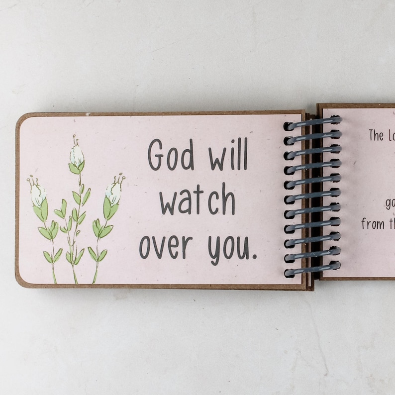 May include: Open spiral-bound journal with a light pink page featuring the text "God will watch over you." The page has a floral illustration in green and white. The journal is open to the first page.