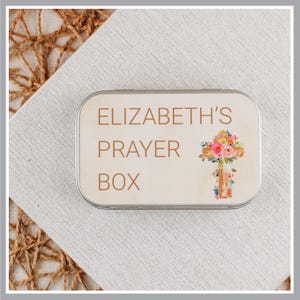 May include: A rectangular metal tin with rounded corners featuring the text ELIZABETH'S PRAYER BOX in brown lettering. To the right of the text is a graphic of a wooden cross adorned with colorful pink, orange, and green floral illustrations.