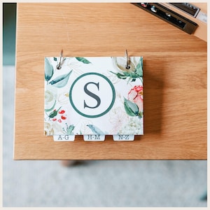 May include: A rectangular address book featuring a white cover with a watercolor floral pattern in shades of green, red, and cream. A large dark green circle contains the letter S in the center. Three index tabs at the bottom are labeled A-G, H-M, and N-Z.