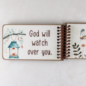 May include: Open wooden journal with a spiral binding. The left page features the text "God will watch over you" with a watercolor illustration of a lantern and foliage. The right page has a butterfly and floral design.