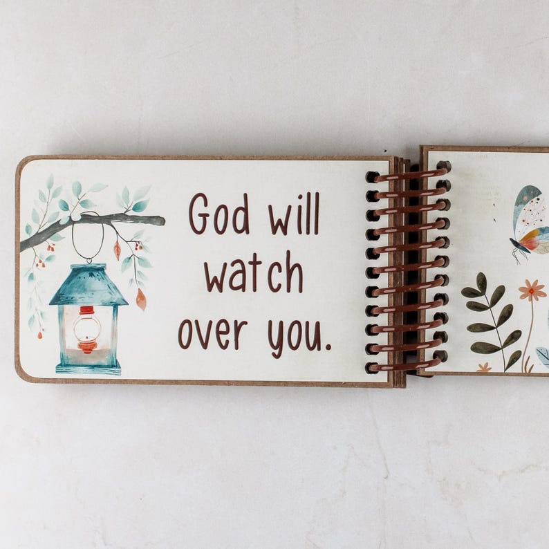 May include: Open wooden journal with a spiral binding. The left page features the text "God will watch over you" with a watercolor illustration of a lantern and foliage. The right page has a butterfly and floral design.