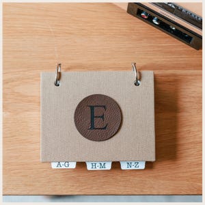 May include: A rectangular beige fabric-covered index card holder with two silver metal rings at the top. A circular dark brown faux leather patch with the letter E is centered on the front. Three white index tabs labeled A-G, H-M, and N-Z.