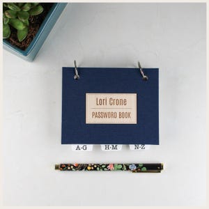 May include: A dark blue password book with a light wood-toned label reading Lori Crone PASSWORD BOOK, featuring three white alphabetical index tabs labeled A-G, H-M, and N-Z, held together by two silver metal rings, accompanied by a floral print pen.