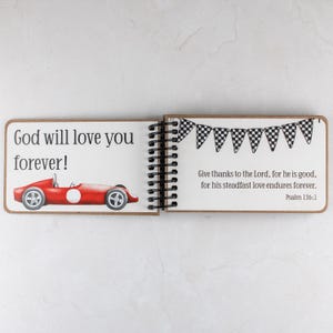 May include: Open spiral-bound journal with a red race car illustration and the text "God will love you forever!". The right page has a banner of checkered flags and a Bible verse. The journal is open on a white surface.
