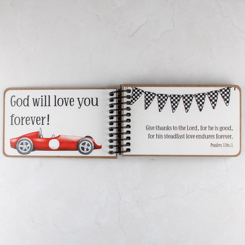 May include: Open spiral-bound journal with a red race car illustration and the text "God will love you forever!". The right page has a banner of checkered flags and a Bible verse. The journal is open on a white surface.