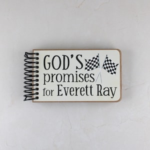 May include: A spiral-bound notebook with a cream-colored cover. The cover features the text "GOD'S promises for Everett Ray" in black, along with two checkered flags. The notebook's binding is black.