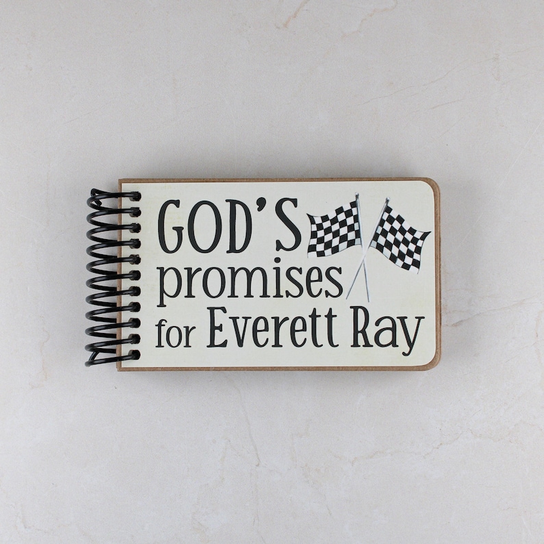 May include: A spiral-bound notebook with a cream-colored cover. The cover features the text "GOD'S promises for Everett Ray" in black, along with two checkered flags. The notebook's binding is black.