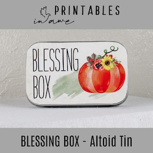 Puede incluir: Una lata de metal blanca con las palabras "Blessing Box" impresas en ella. La lata tiene una imagen de acuarela de una calabaza con flores y hojas.