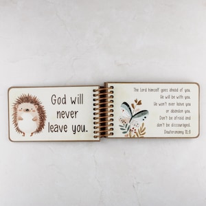 May include: Open wooden book with spiral binding, featuring a cartoon hedgehog and a butterfly illustration. The left page reads "God will never leave you." The right page includes a Bible verse from Deuteronomy 31:8.