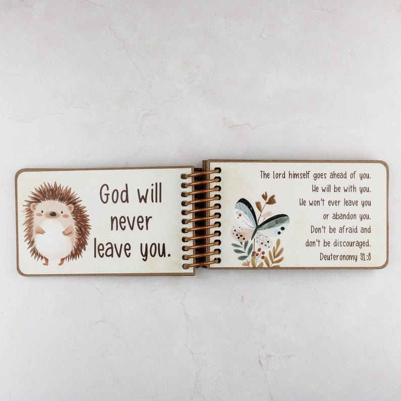 May include: Open wooden book with spiral binding, featuring a cartoon hedgehog and a butterfly illustration. The left page reads "God will never leave you." The right page includes a Bible verse from Deuteronomy 31:8.
