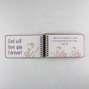 May include: Open spiral-bound journal with the text "God will love you forever!" on the left page. The right page reads "Thank the lord, because he is good! For his trustworthy love lasts forever. Psalm 136:1." Floral illustrations are present.