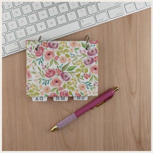 May include: A small ring-bound address book featuring a floral pattern with pink, purple, and green watercolor-style flowers and leaves. The book includes three white index tabs labeled A-G, H-M, and N-Z. A pink retractable pen is placed nearby.