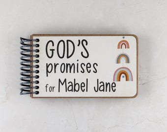 Puede incluir: Un cuaderno de espiral con una cubierta blanca que presenta el texto negro "GOD'S promises for Mabel Jane" y una pila vertical de tres ilustraciones de arcoíris de tonos apagados a la derecha, encuadernado con una espiral de metal negro.