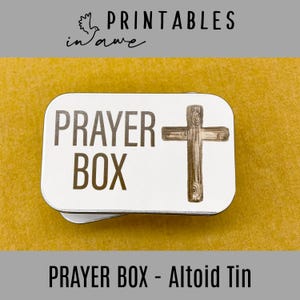 May include: A white metal tin with the words "PRAYER BOX" and a wooden cross graphic. The tin is on a yellow surface.