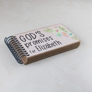 May include: A small, rectangular notebook with a spiral binding. The cover is light pink with the text "GOD'S promises for Elizabeth" in black. Floral accents in pastel colors decorate the upper right corner. The pages are a light brown color.
