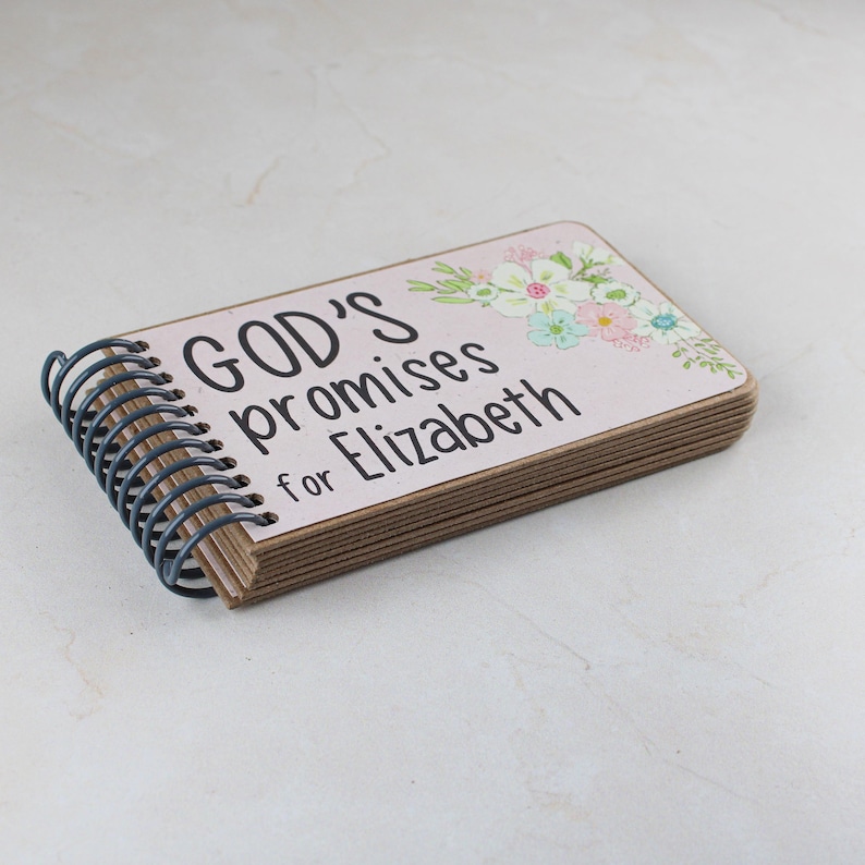 May include: A small, rectangular notebook with a spiral binding. The cover is light pink with the text "GOD'S promises for Elizabeth" in black. Floral accents in pastel colors decorate the upper right corner. The pages are a light brown color.