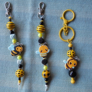 May include: Three bee-themed keychains. Each keychain features a silver clasp, yellow and black striped beads, and a cartoon bee charm. Additional beads are black, light blue, and yellow. One keychain has a yellow ring.