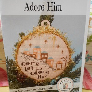 May include: A cross-stitch pattern booklet titled Adore Him featuring a circular ornament design with a nativity scene, gold tinsel trim, and the text Come let us adore Him. The cover displays a finished needlework project on a pine branch.