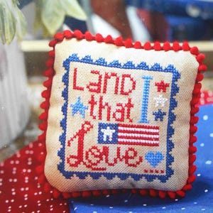 May include: A square beige fabric pillow with red pom-pom trim featuring a cross-stitched design. The text reads Land that I Love in red and blue. It includes a small American flag graphic, a blue star, a white star, and a blue heart.