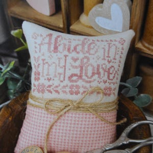 May include: A small, decorative pillow with the embroidered text "Abide in My Love" in pink. The pillow is tied with twine and has a pink and white checkered pattern on the bottom half. A pair of scissors rests in front.