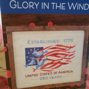May include: A cross-stitch pattern titled GLORY IN THE WIND featuring a blue square with white stars, three light blue birds, and red and white wavy stripes. The text reads ESTABLISHED 1776, UNITED STATES OF AMERICA, and 250 YEARS in a wooden frame.