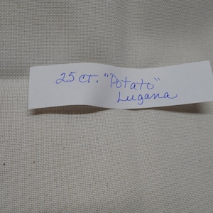 May include: A piece of off-white fabric with a label that reads "25 ct. 'Potato' Lugana" in blue ink. The fabric has a visible cross-stitch pattern.