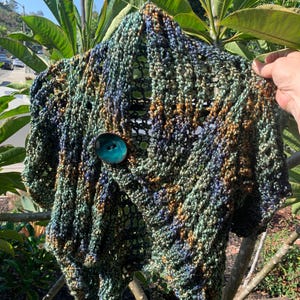Op de afbeelding: Een handgebreid vestje van gemêleerd garen in tinten bosgroen, marineblauw en metallic brons. Het kledingstuk heeft een open, grofgebreide textuur en wordt aan de voorzijde gesloten met een enkele, grote, ronde, blauwgroene knoop.