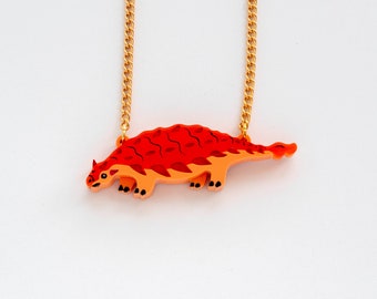 May include: A gold chain necklace with a red and orange dinosaur pendant. The dinosaur is stylised and has a scaly texture.