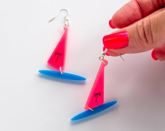 May include: A pair of pink and blue sailboat earrings with a palm tree design. The earrings are made of plastic and have silver hooks.