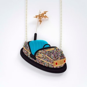 May include: A glittery black bumper car necklace with a blue interior and a gold star on top. The necklace is on a silver chain.