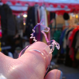 Op de afbeelding: Een transparante plastic ring met een unicorn silhouet ontwerp. De ring wordt gedragen aan een vinger.