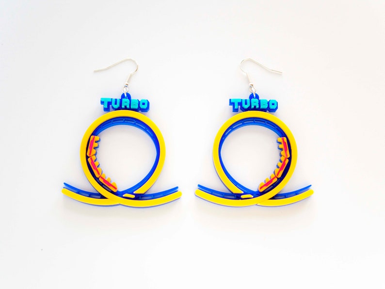Pu&ograve; includere: Un paio di orecchini blu e gialli a forma di anello di montagne russe con la scritta "TURBO" in cima.