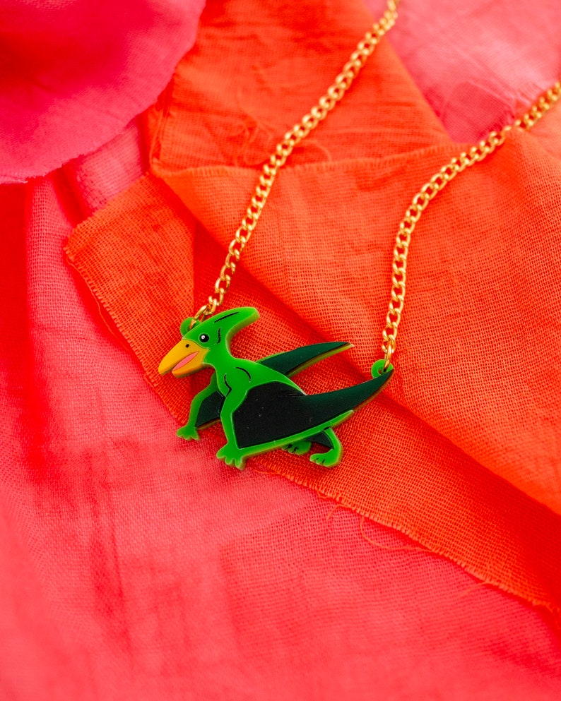 May include: A gold chain necklace with a green and black pterodactyl pendant. The pendant is made of plastic and has a cartoon-like design.