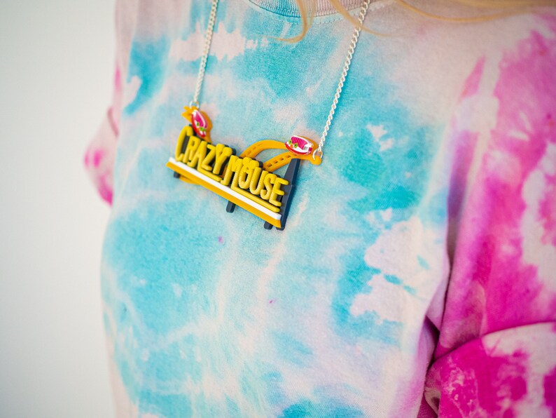May include: A yellow and black necklace with the words "Crazy Mouse" on it. The necklace is worn by a person wearing a tie-dye shirt.