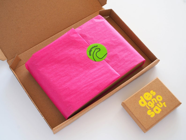 Puede incluir: Una caja de cart&oacute;n marr&oacute;n con un art&iacute;culo envuelto en papel de seda rosa en su interior. La caja tiene una pegatina verde con una ilustraci&oacute;n de dinosaurio y el texto "designonour". Tambi&eacute;n se ve una caja marr&oacute;n m&aacute;s peque&ntilde;a con el texto "des 1920 sauv" en amarillo.