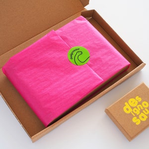 May include: A brown cardboard box with a pink tissue paper-wrapped item inside. The box has a green sticker with a dinosaur illustration and the text "designonour". A smaller brown box with the text "des 1920 sauv" in yellow is also visible.